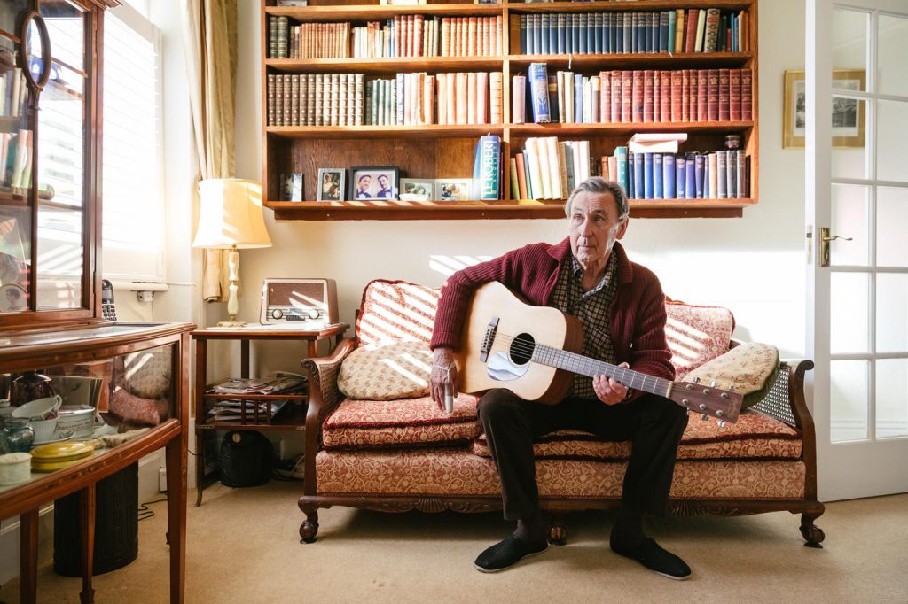 Jay Dowle playing an acoustic guitar on the sofa in a room lined with books, light and sunshine.